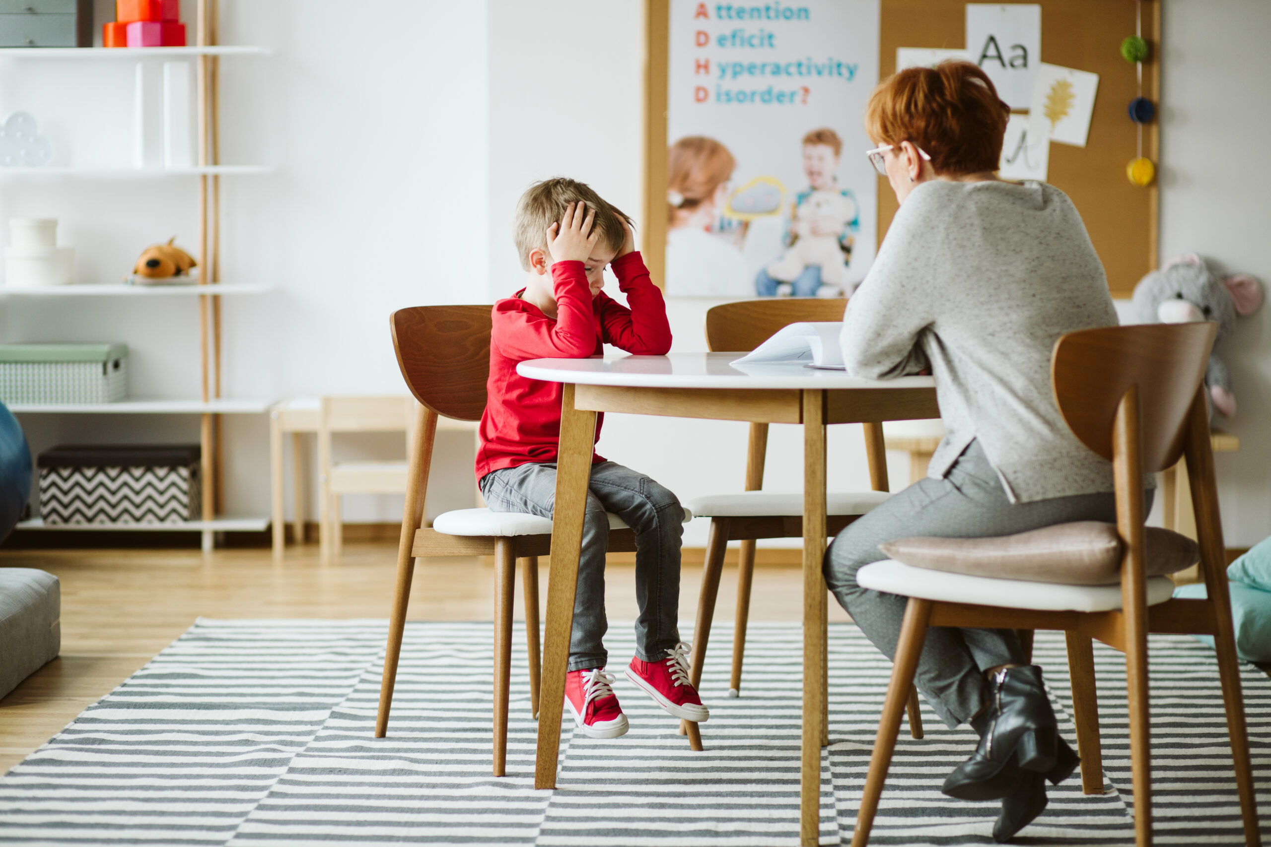 A little boy sits across from an adult at a table. He has his hands on his head as if frustrated or bored.