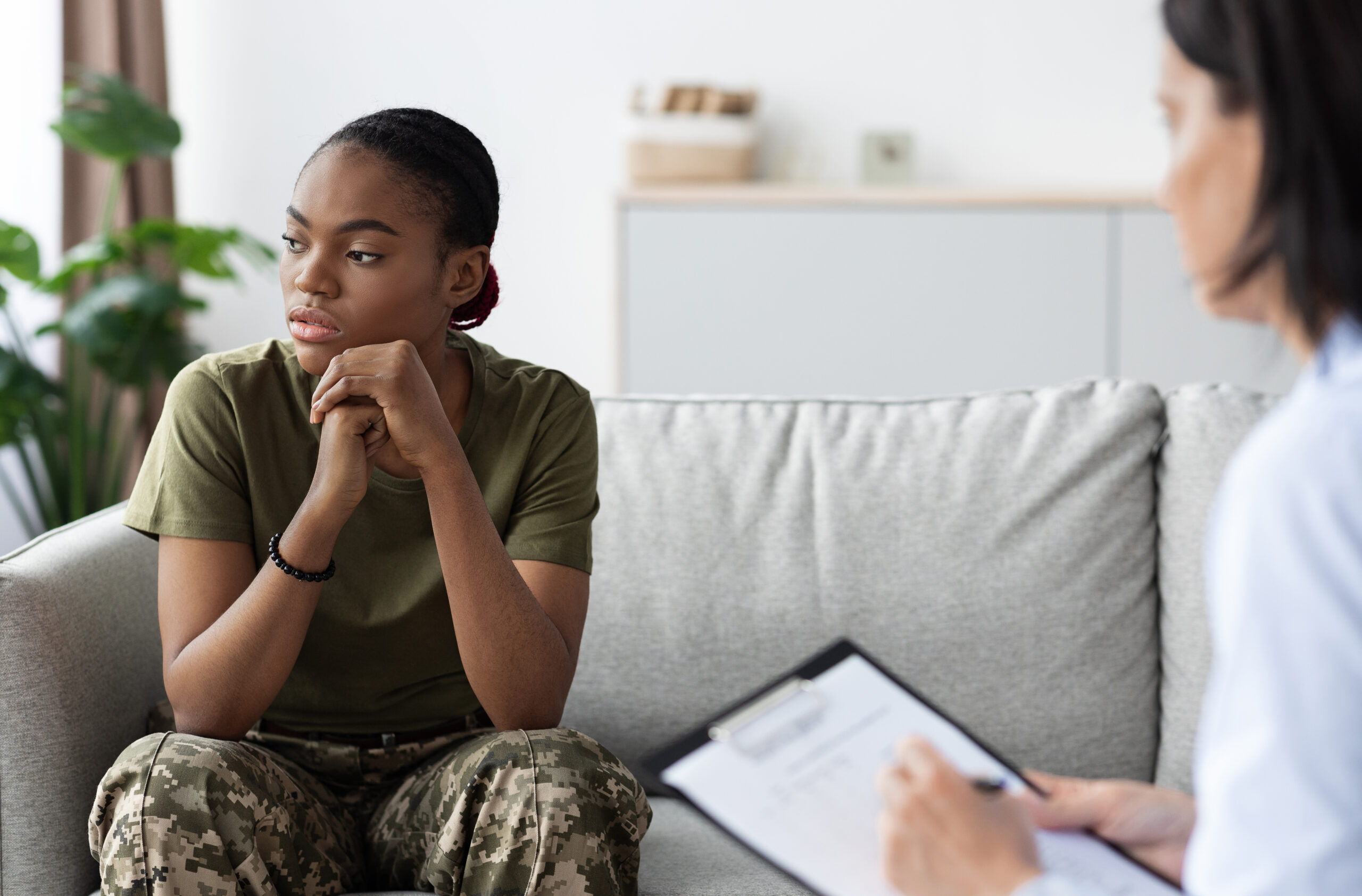 A woman in army clothes sits on a couch, with her chin resting on her hands. Her gaze is to the side, appearing in deep thought. In front of her a therapist sits with a clipboard, writing notes.