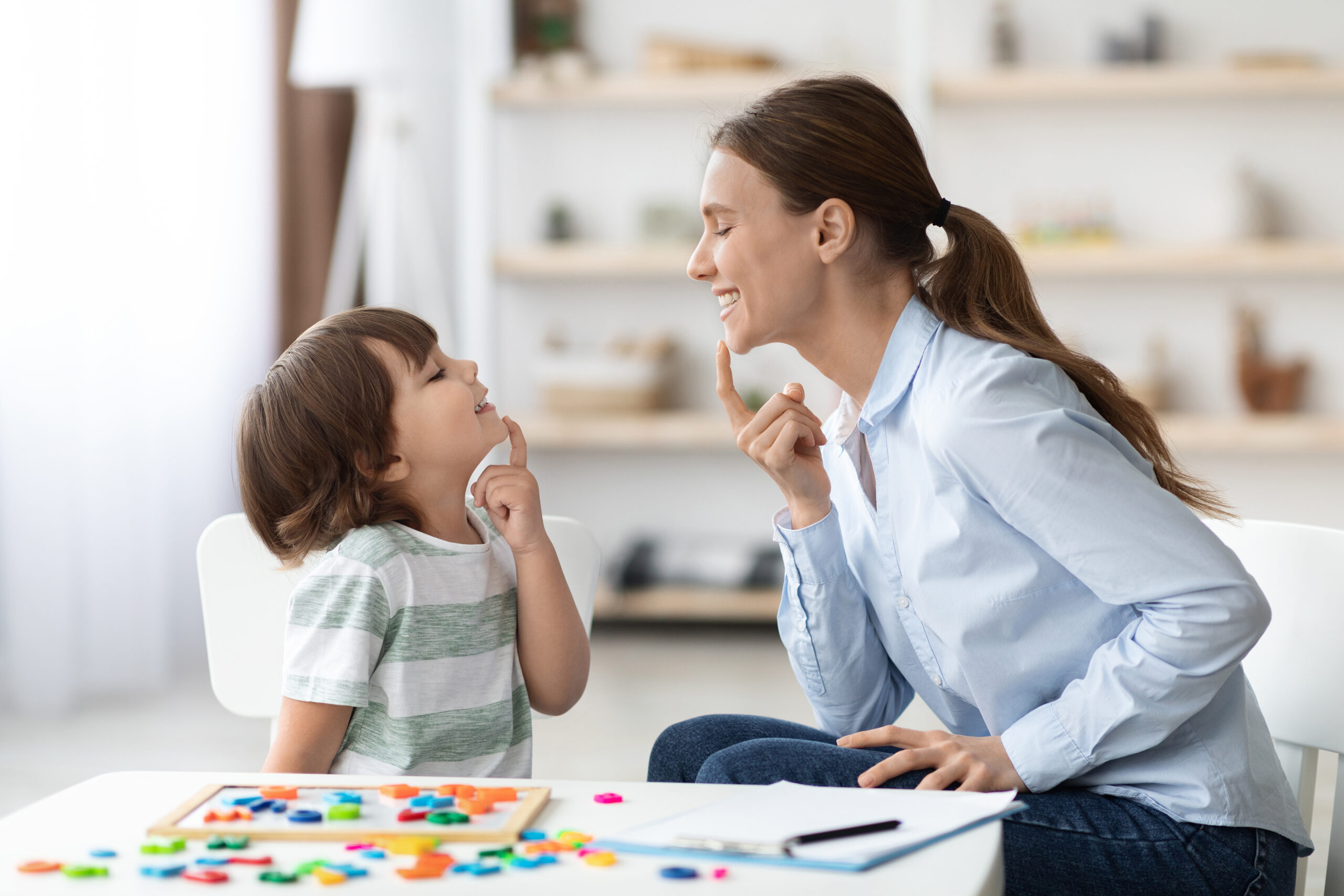 A young boy and a woman sit at a small table facing each other. They are smiling and communicating nonverbally by both touching their chin. On the table sits a puzzle and a clipboard with a pen sitting on top of it.
