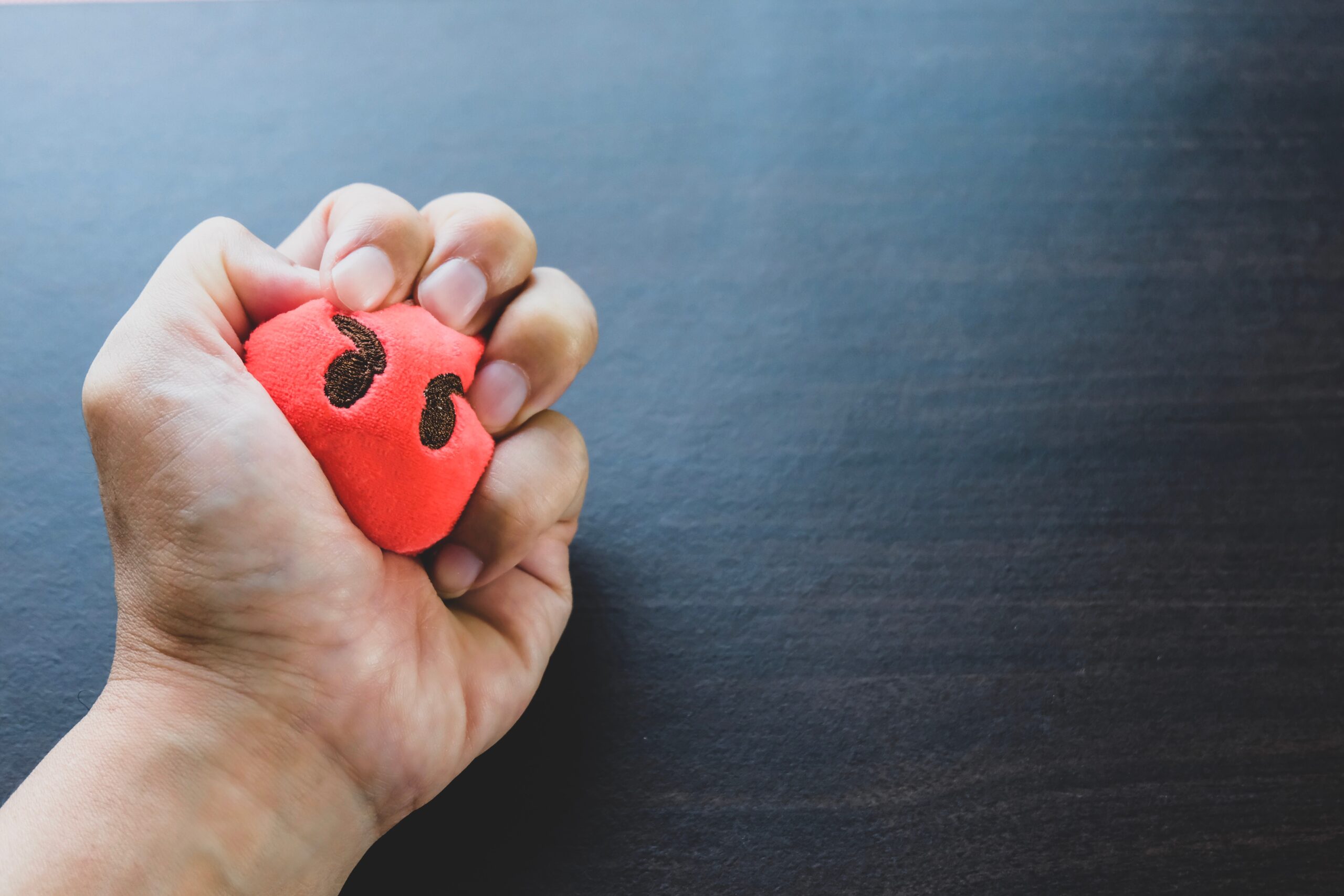 A man’s hand is gripping a red stress ball with an angry face, seemingly using it as a coping mechanism for anger.
